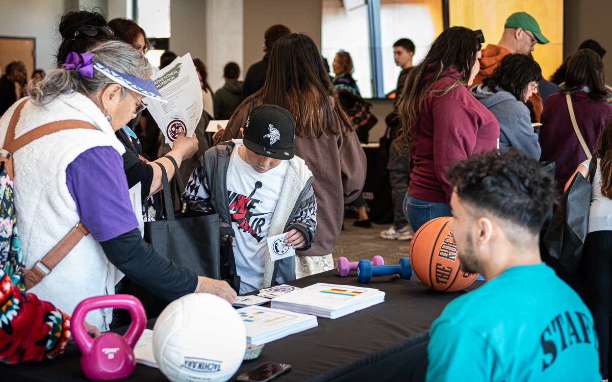 A Native Fitness and Nutrition staffer sits at a black table that has a basketball, volleyball, and weight on it, and talks to visitors about programming.