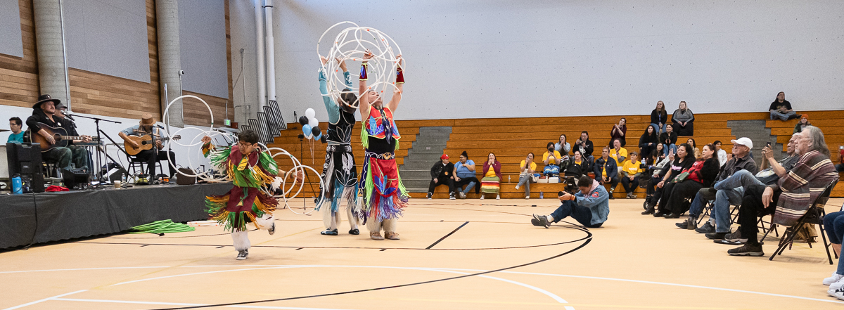 The Sampson Brothers hoop dance in the Minneapolis American Indian Center gym. They wear traditional regalia and a group of attendees sit in chairs in the gym to watch.