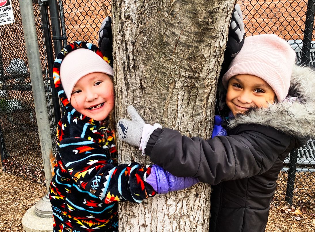 Two elementary school-aged girls in the Boys and Girls Club smile at the camera as they hug a tree.