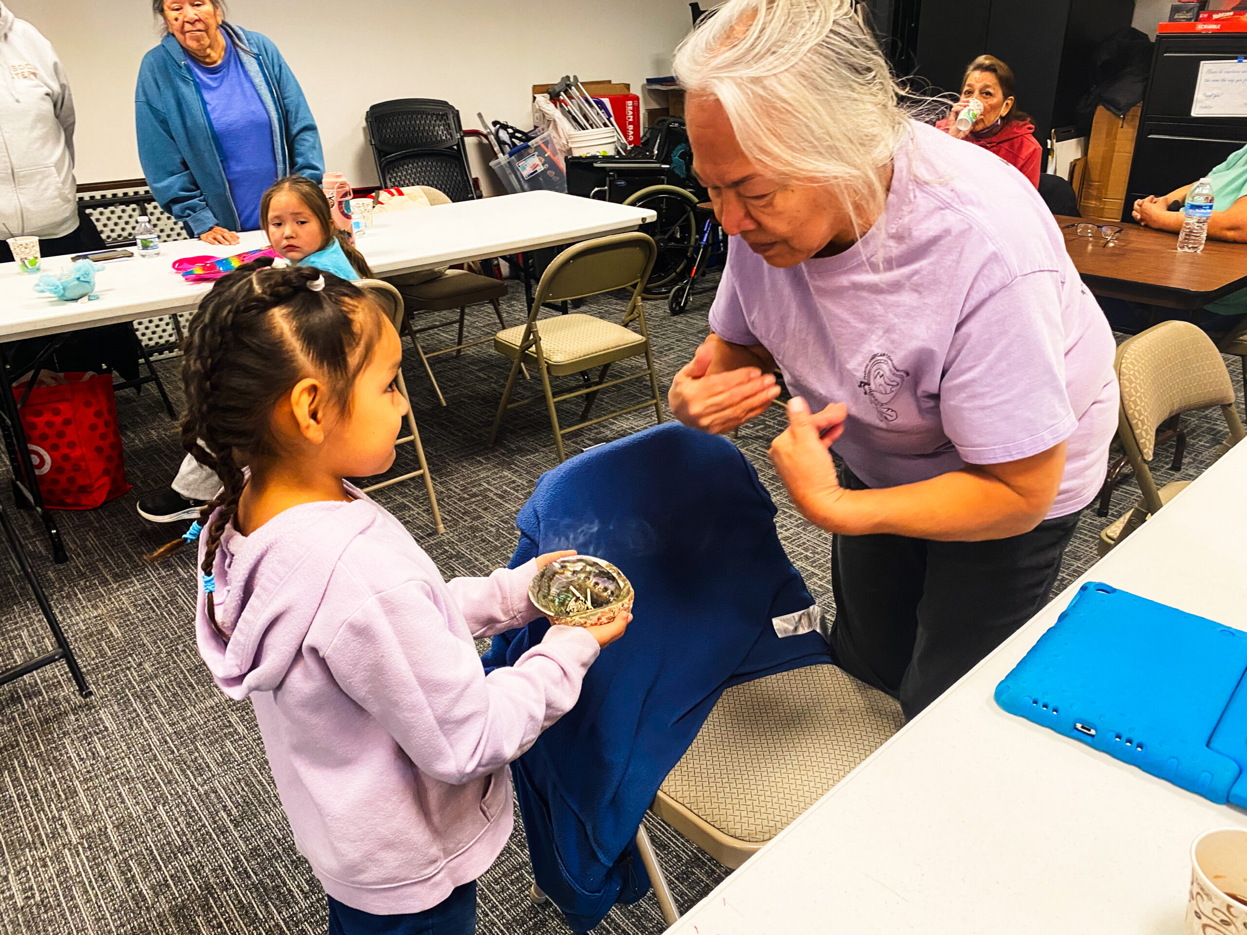 A young Native girl shows a Native elder woman a bowl of cedar, tobacco, sage, and sweetgrass.