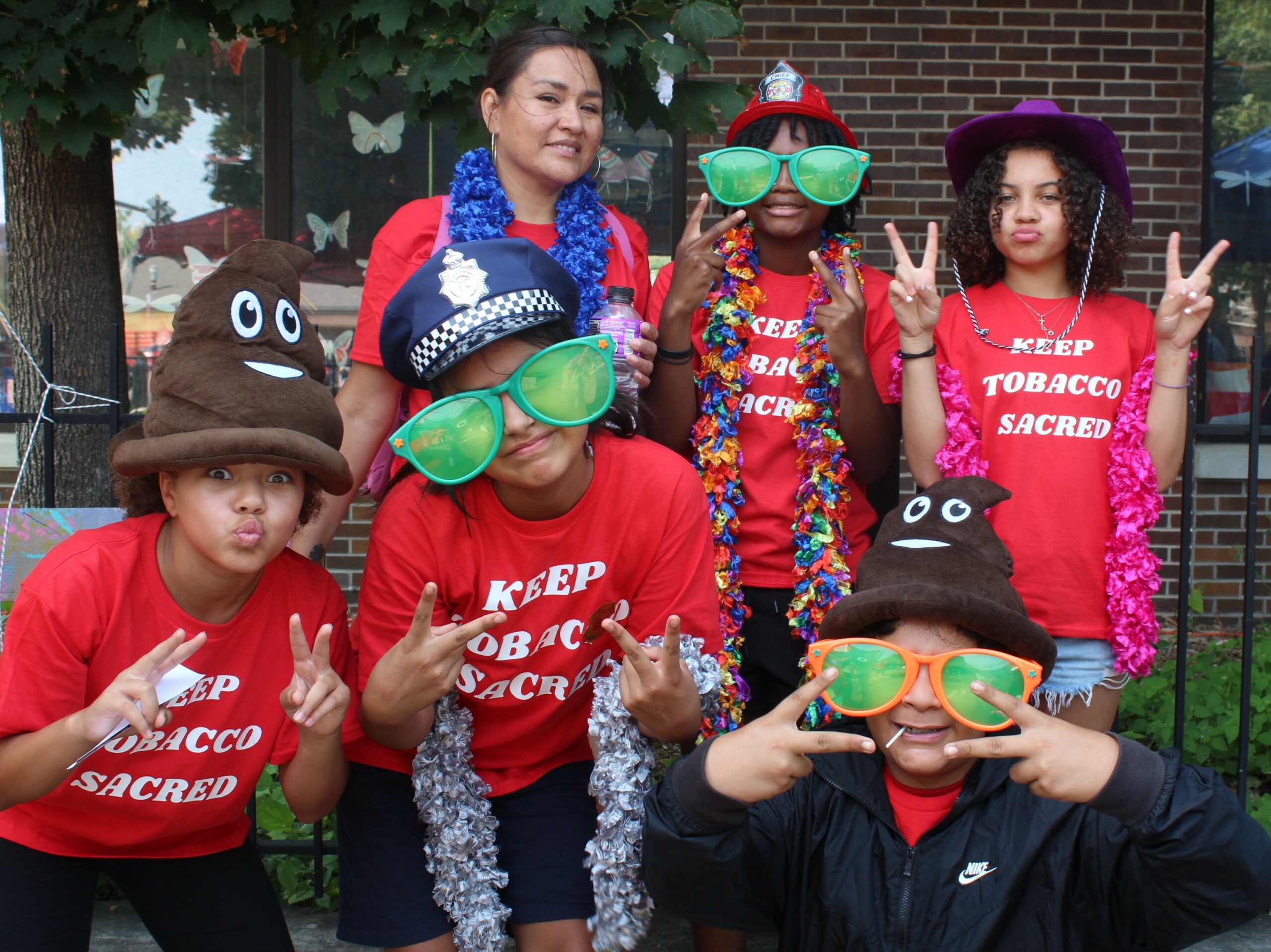 A group of 6 Native teenagers wear read tshirts and wear costume hats and large sunglasses.